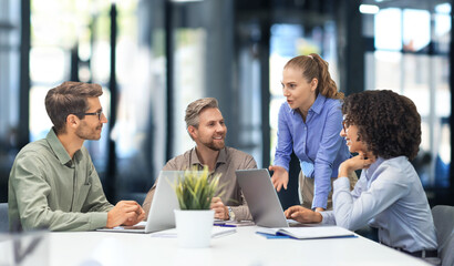 Vier glücklich aussehende Menschen in einer Geschäftsbesprechung. Eine Frau steht, eine andere Frau und zwei Männer sitzen dafür im Kreis an einem Tisch mit Laptops vor sich.Foto: (c) ty / AdobeStock