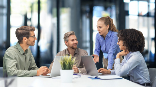 Vier glücklich aussehende Menschen in einer Geschäftsbesprechung. Eine Frau steht, eine andere Frau und zwei Männer sitzen dafür im Kreis an einem Tisch mit Laptops vor sich.Foto: (c) ty / AdobeStock