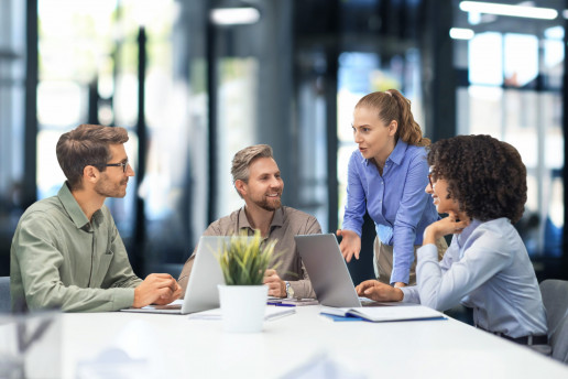 Vier glücklich aussehende Menschen in einer Geschäftsbesprechung. Eine Frau steht, eine andere Frau und zwei Männer sitzen dafür im Kreis an einem Tisch mit Laptops vor sich.Foto: (c) ty / AdobeStock